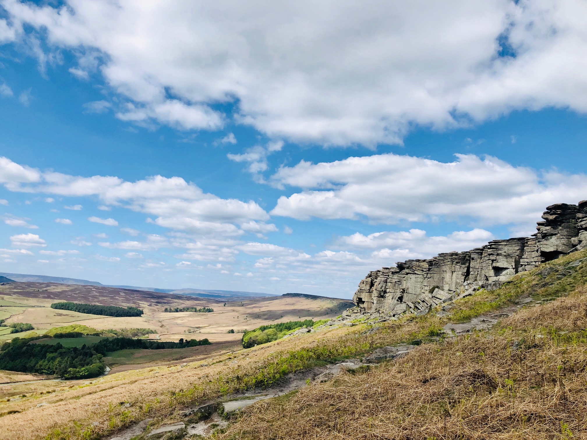 Rolling Derbyshire moorlands leading up to the dramatic gritstone cliffs of Stanage Edge, bathed in soft daylight.